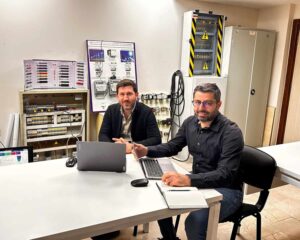 Açıklama: Elektrik tesisatı ve kontrol panelleri üzerinde çalışan iki elektrik teknisyeni, teknik ekipmanlar ve diyagramlar ile opti odaklı çalışma ortamında.